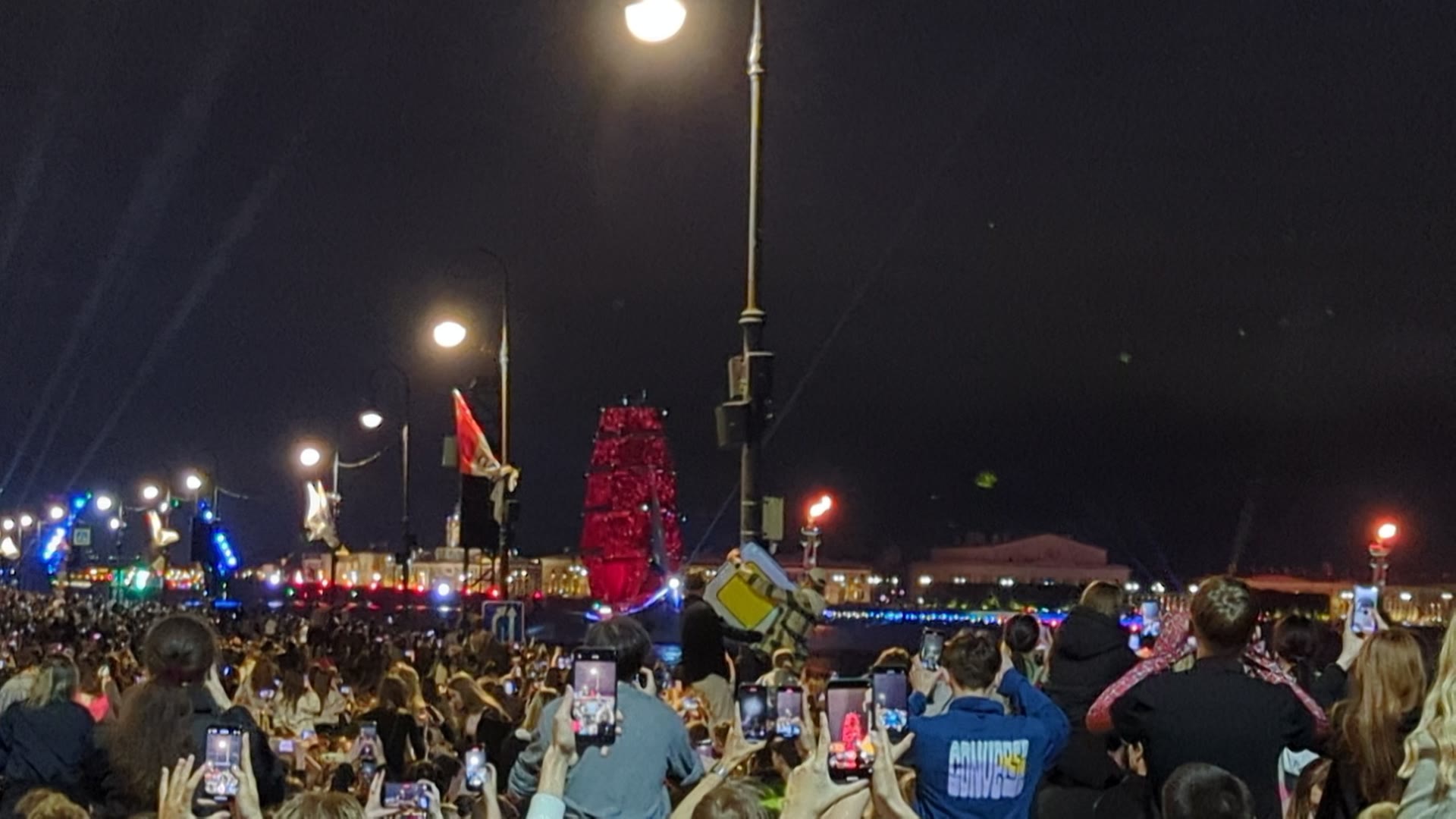 a crowd on the neva embankment at night, phones raised, the scarlet-sailed brig lit red on the water