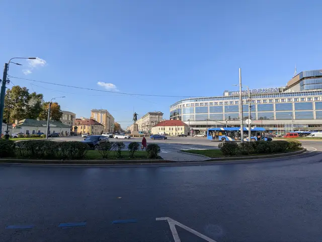 a wide square around an equestrian statue, a curved glass hotel and a blue trolleybus behind it