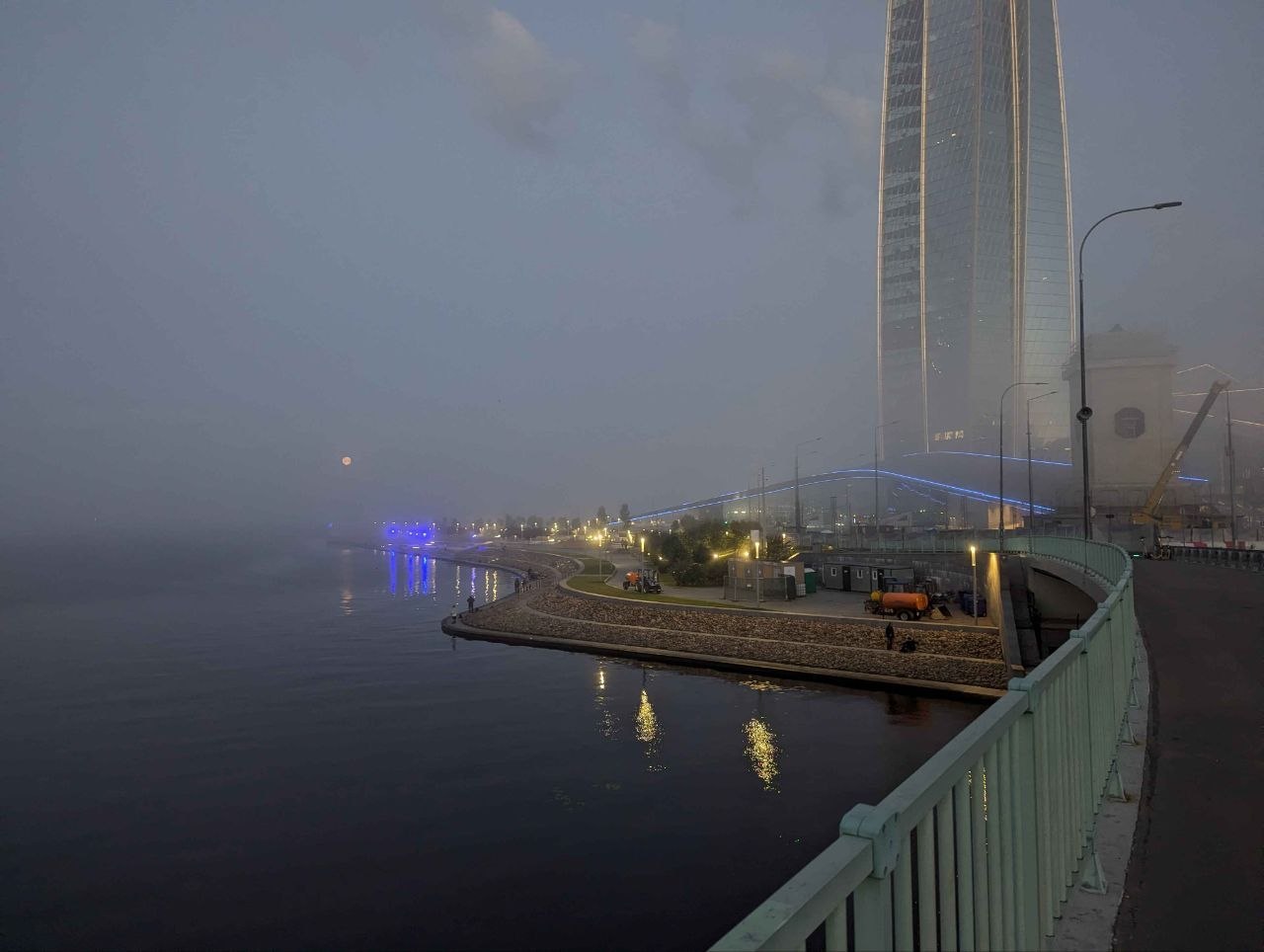 a glass tower disappearing into fog at blue hour, embankment lamps doubled in the water below