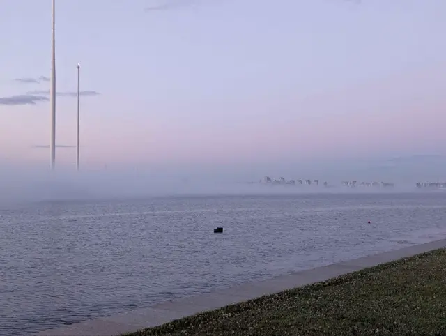 fog lying flat across the gulf at dusk, the far shore's towers only just breaking through it