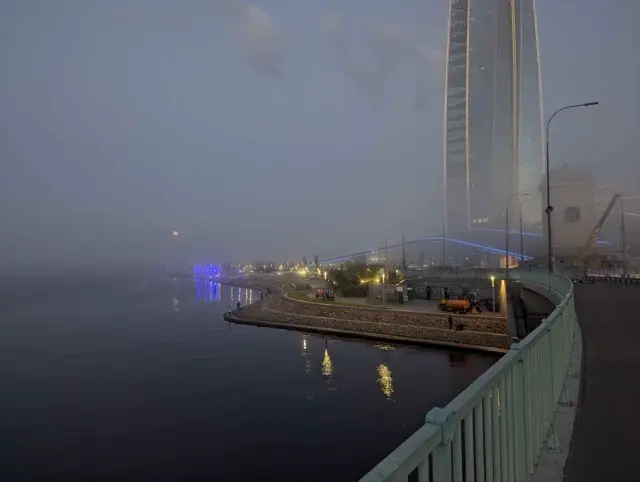 a glass tower disappearing into fog at blue hour, embankment lamps doubled in the water below