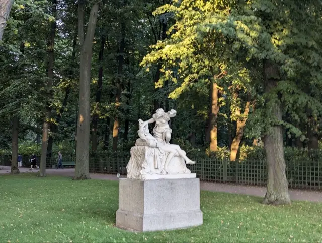 a white marble group on a granite pedestal in a park, evening sun catching the leaves behind it