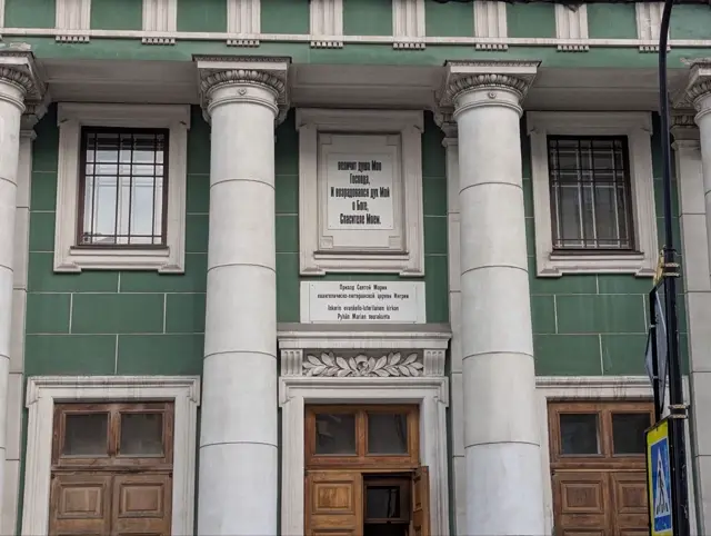 a green facade with white columns, a verse painted above the door and a bilingual russian and finnish plaque below it